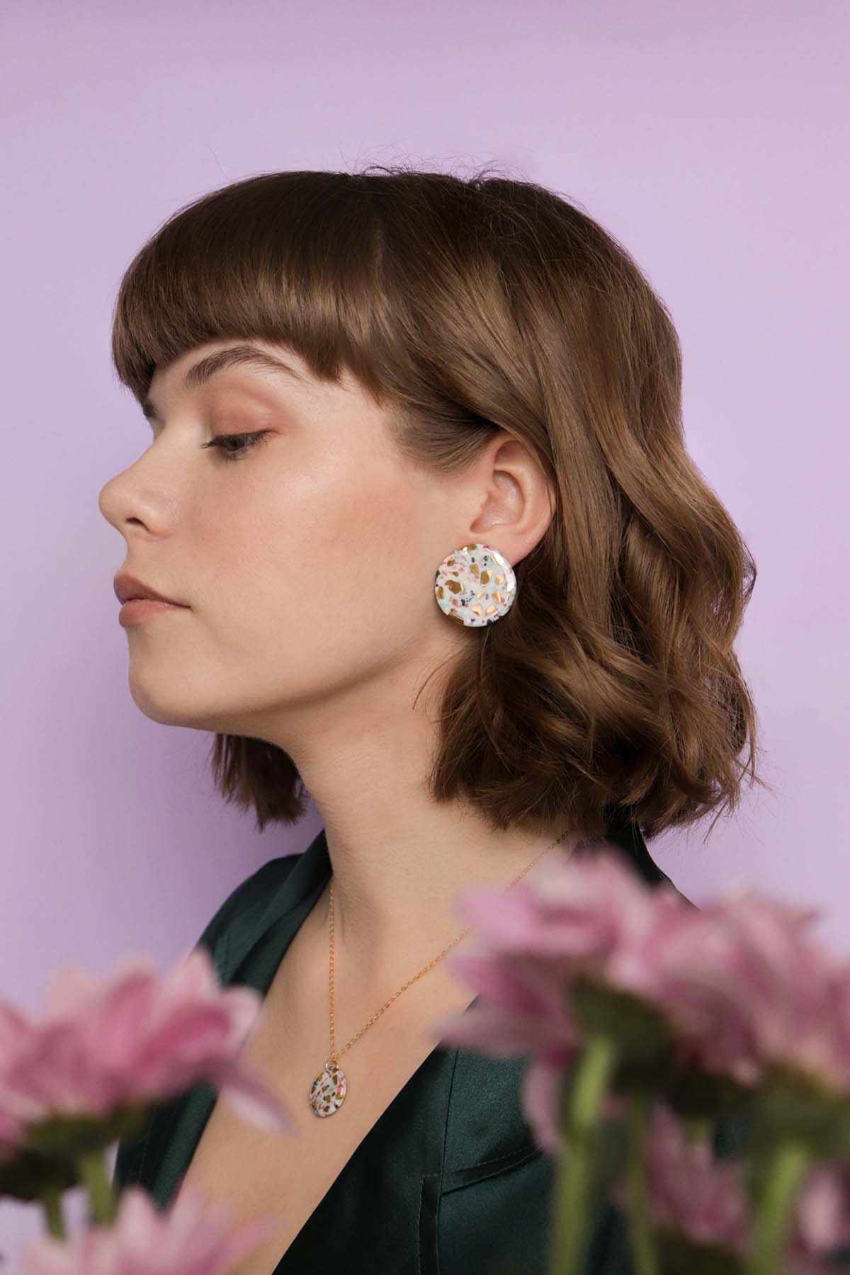A woman with short brown hair wears a dark green top and Rozenthal Jewelrys Astilla Recycled Studs / M, posing in profile against a light purple background with pink flowers in the foreground.