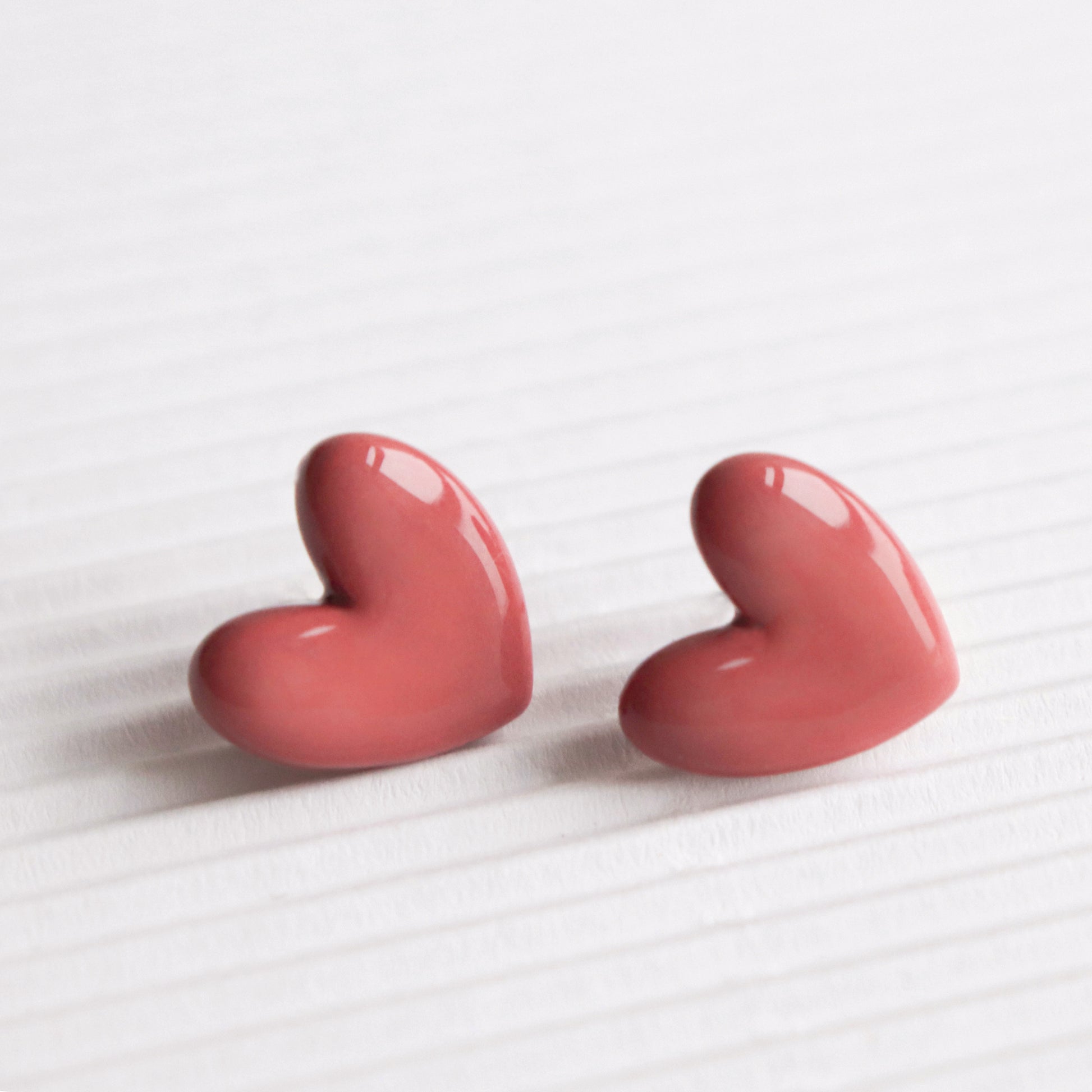 Two glossy, pink heart-shaped Heart Studs by Rozenthal Jewelry rest side by side on a white textured surface with horizontal lines. Handmade in Latvia, these hypoallergenic designer earrings are slightly angled toward each other.