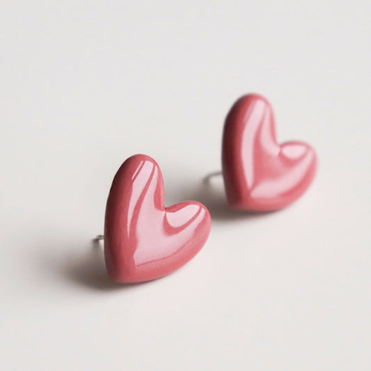 The Heart Studs by Rozenthal Jewelry are two pink, heart-shaped earrings displayed on a white surface. Handmade in Latvia, these lightweight, waterproof studs have a smooth, glossy finish and are shown at slight angles for a 3D effect.