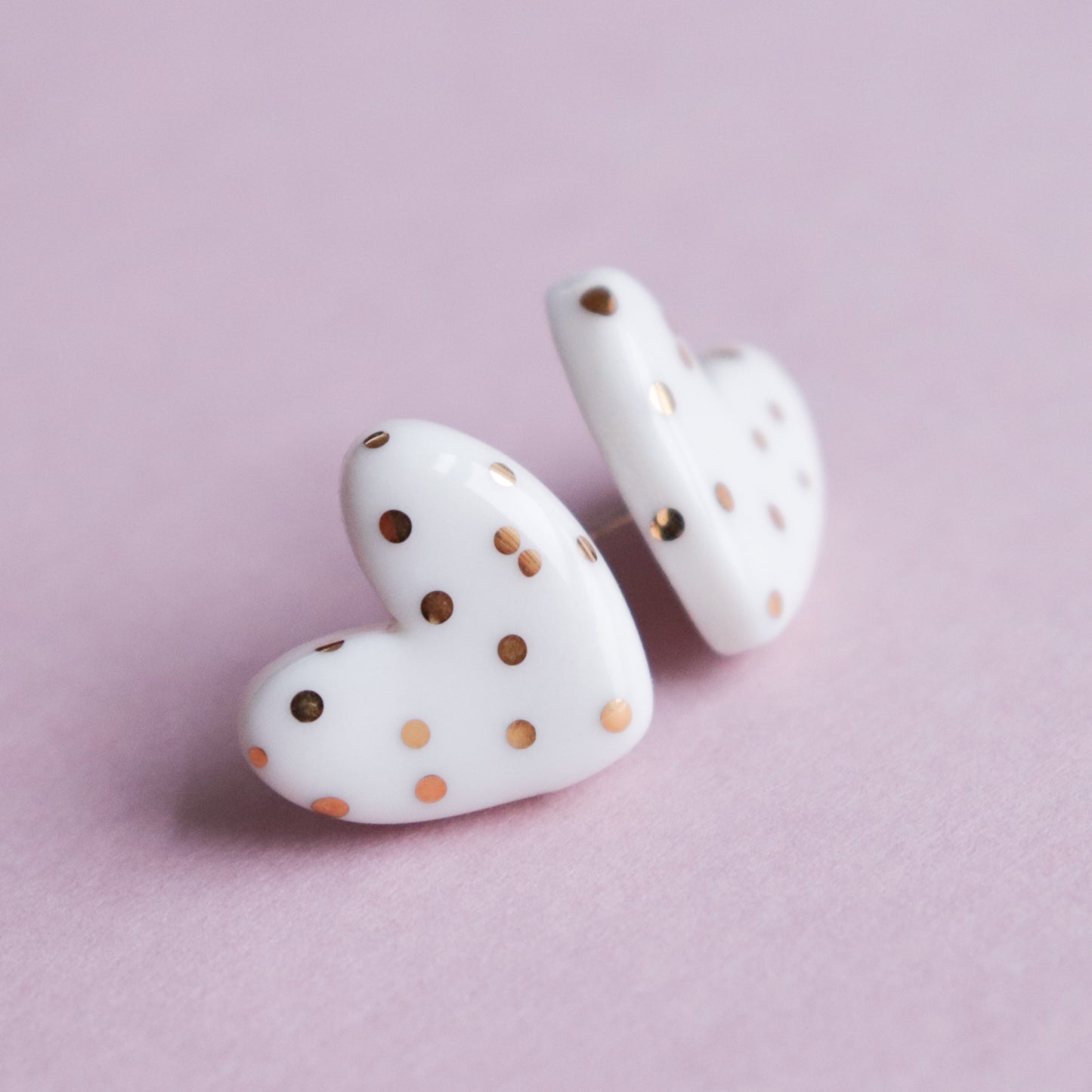 The Heart Studs by Rozenthal Jewelry are white, heart-shaped handmade earrings with gold polka dots, shown on a soft pink surface.