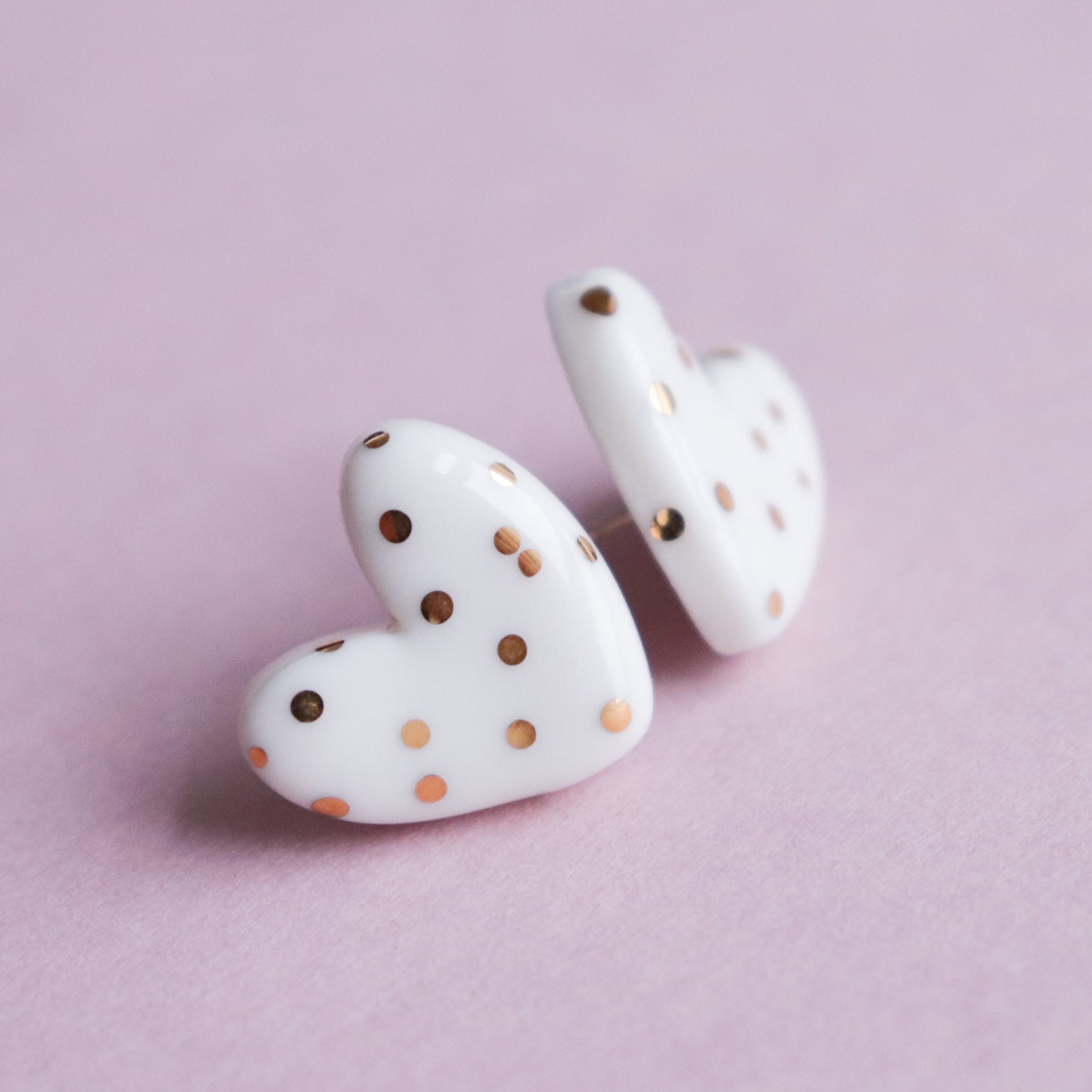 The Heart Studs by Rozenthal Jewelry are white, heart-shaped handmade earrings with gold polka dots, shown on a soft pink surface.