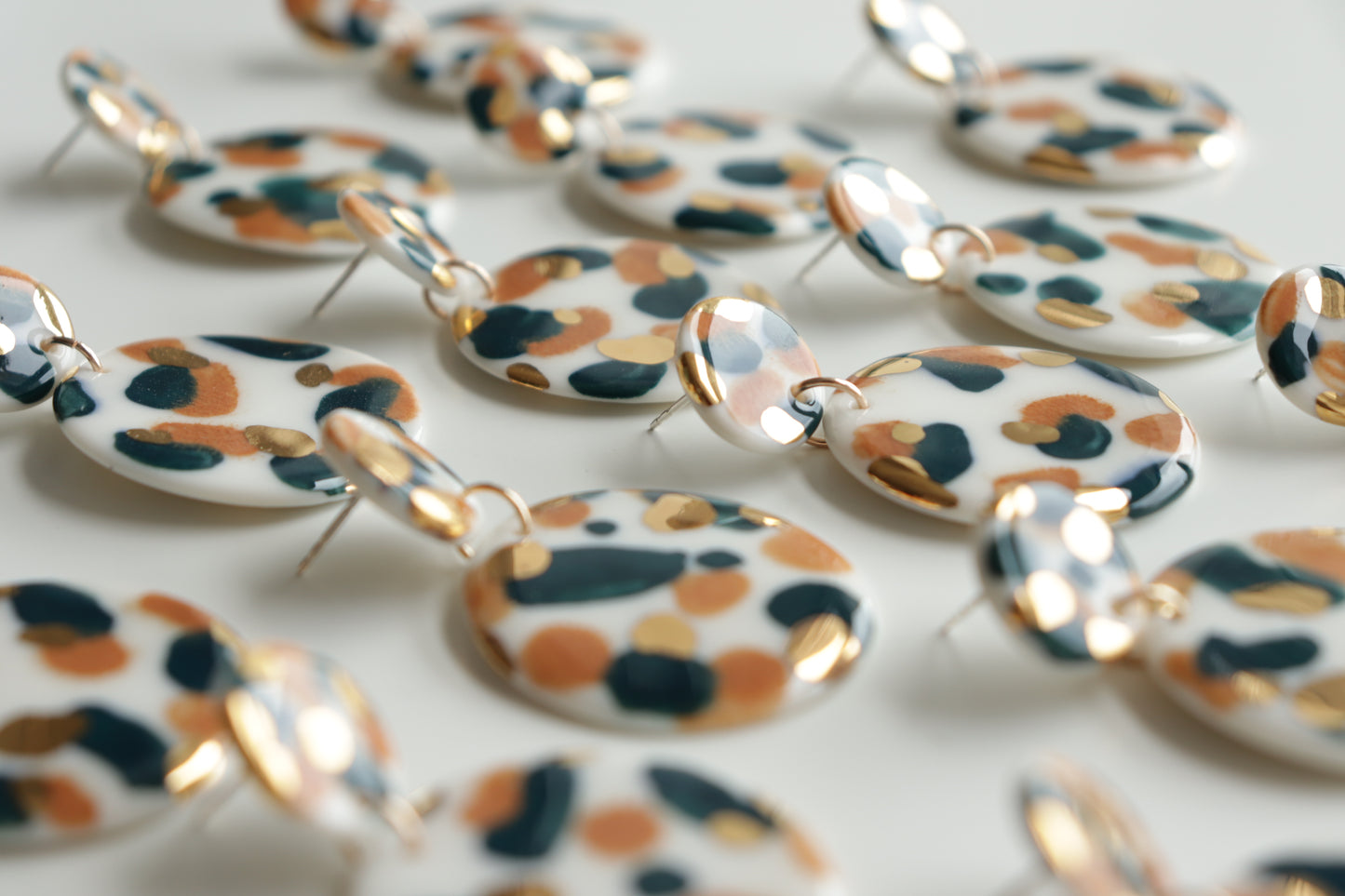 Close-up of several pairs of Rozenthal Jewelry’s Tache Earrings, featuring round porcelain shapes with hand-painted black, brown, and 24K gold leopard spots on a white background, displayed in neat rows.