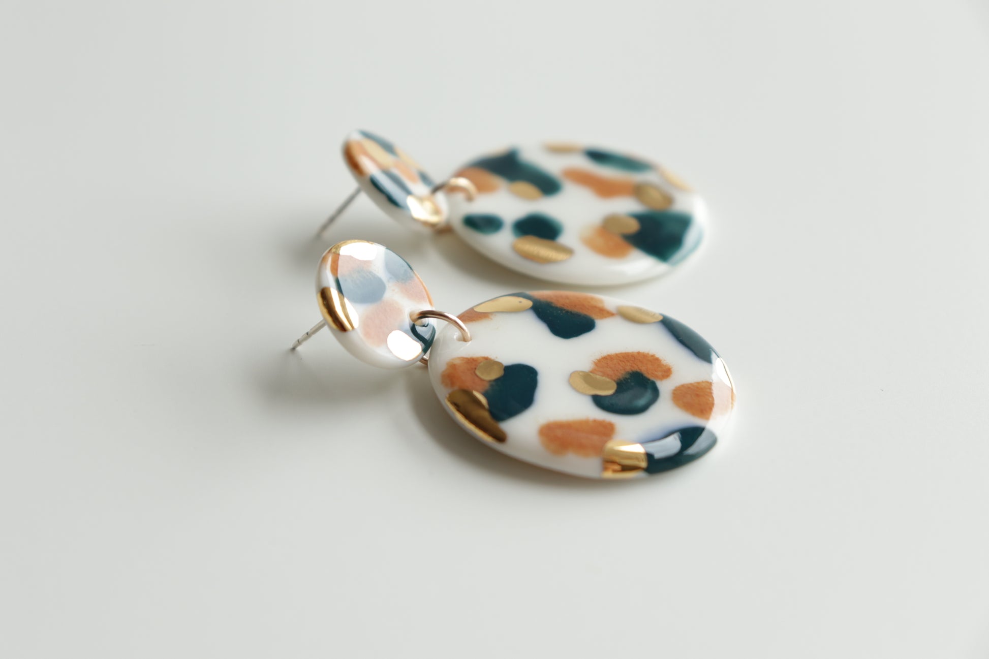 The Tache Earrings by Rozenthal Jewelry are round, dangling porcelain pieces with a glossy finish, featuring an abstract pattern in white, orange, green, and hand-painted 24K gold on a light base, shown on a plain white background.
