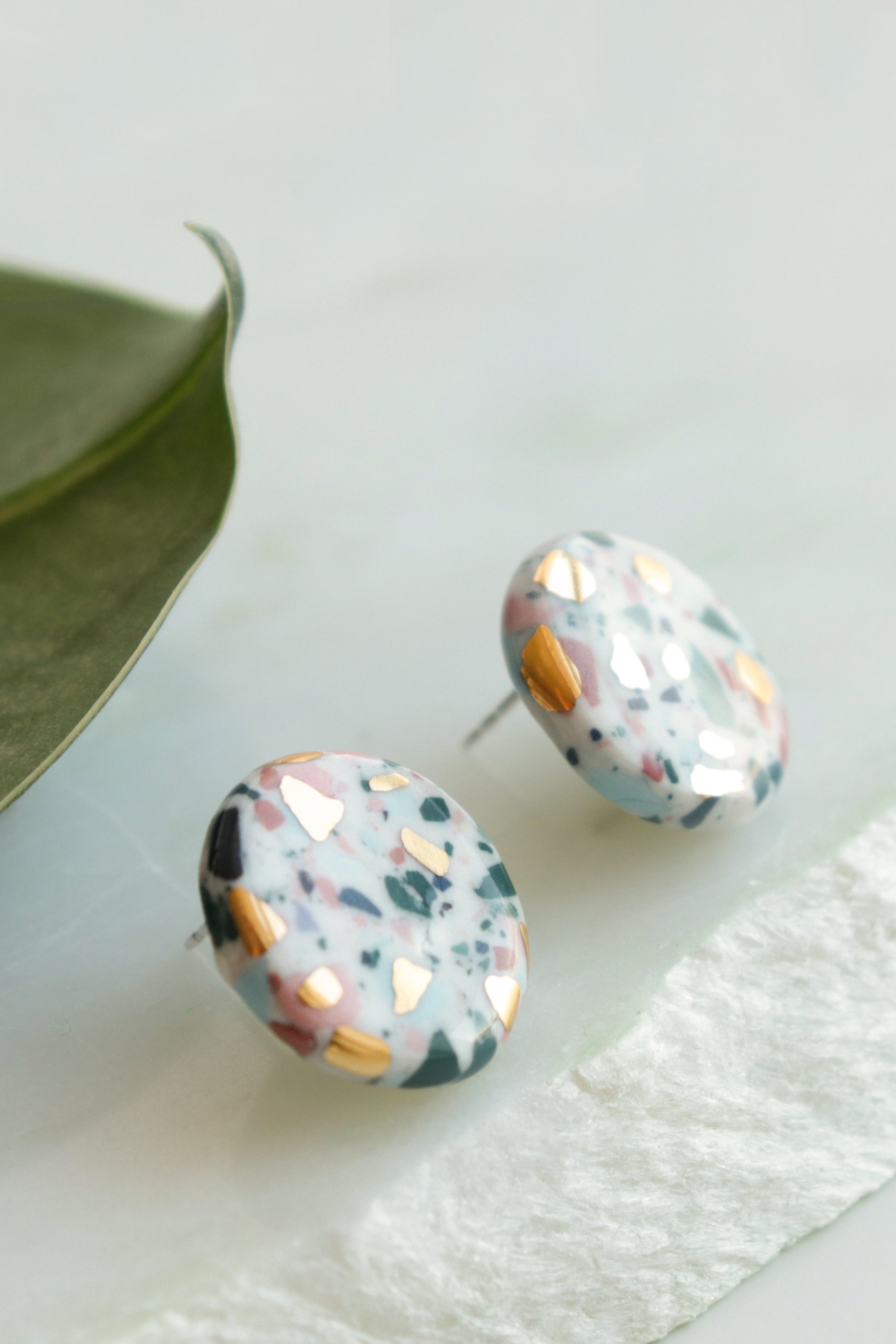 Astilla Recycled Studs / S by Rozenthal Jewelry feature a terrazzo pattern in white, teal, pink, and gold flecks. Eco-friendly and handmade, these unique round studs add charm to any look. Displayed on a light surface beside a green leaf.