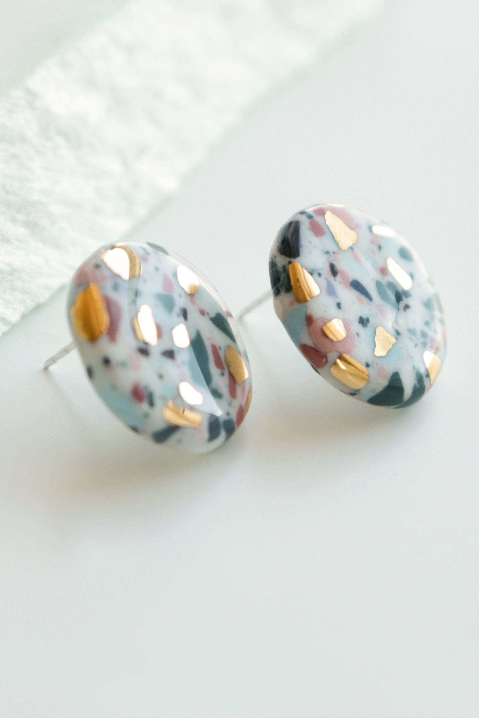 Close-up of Rozenthal Jewelrys Astilla Recycled Studs / S—unique handmade round earrings with a marbled mix of white, black, pink, blue, and gold metallic accents, displayed on a white surface.