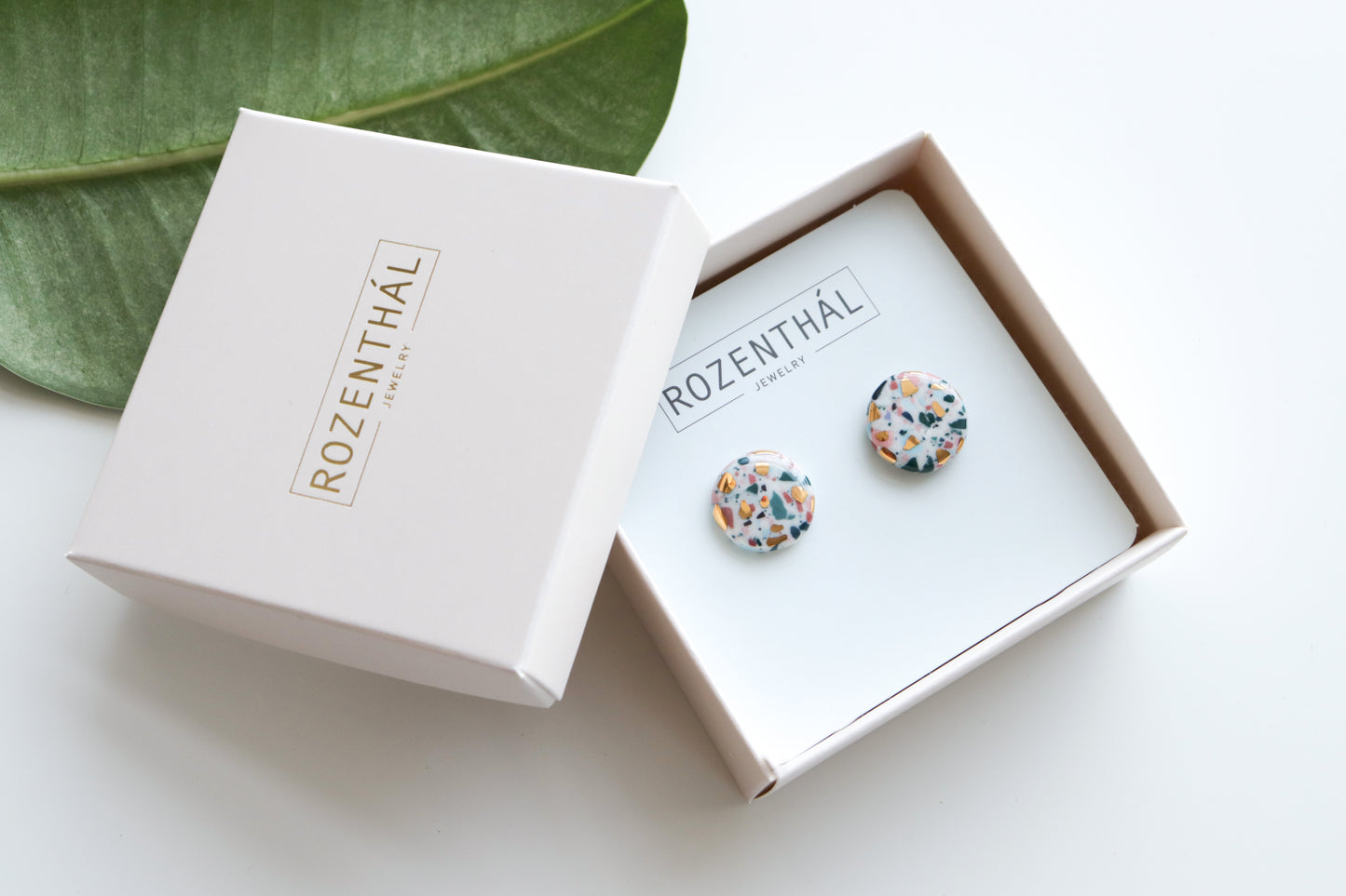 Astilla Recycled Studs / S by Rozenthal Jewelry—colorful, round terrazzo-patterned porcelain earrings—are shown in a white Rozenthal jewelry box, partially open beside a green leaf.