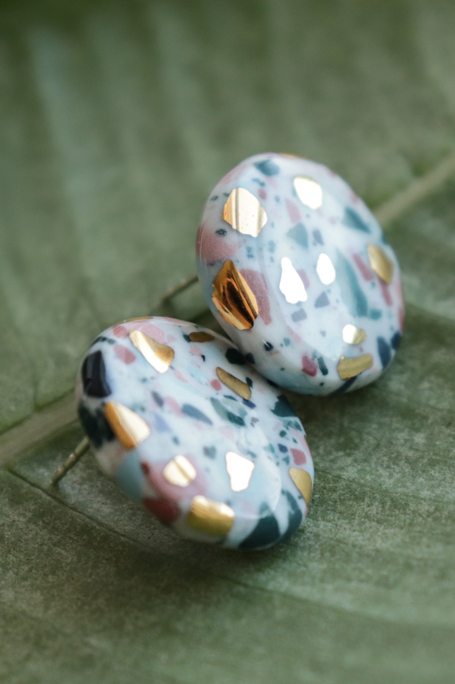 The Astilla Recycled Studs / S by Rozenthal Jewelry are handmade round earrings featuring a marbled pink, blue, green, and white design with gold accents, displayed on a green leaf background.