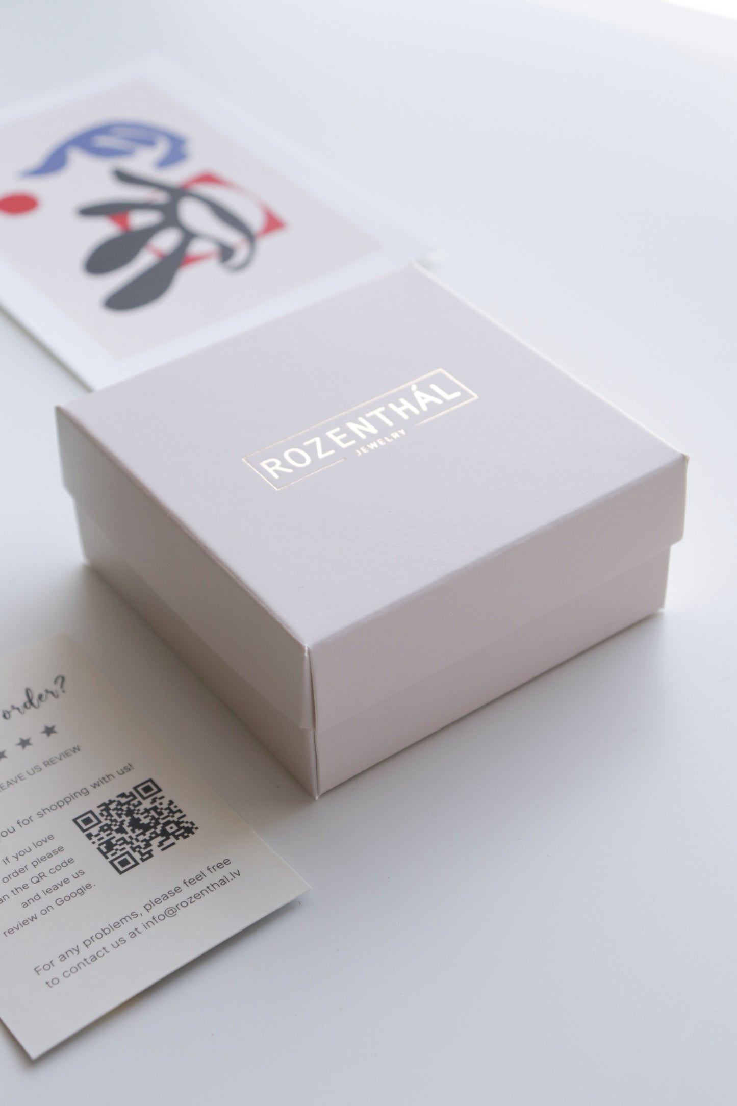 A white ROZENTHAL JEWELRY box sits on a white surface, beside an abstract art card and a QR code business card—ideal packaging for their handmade Drop Earrings / M by Rozenthal Jewelry.
