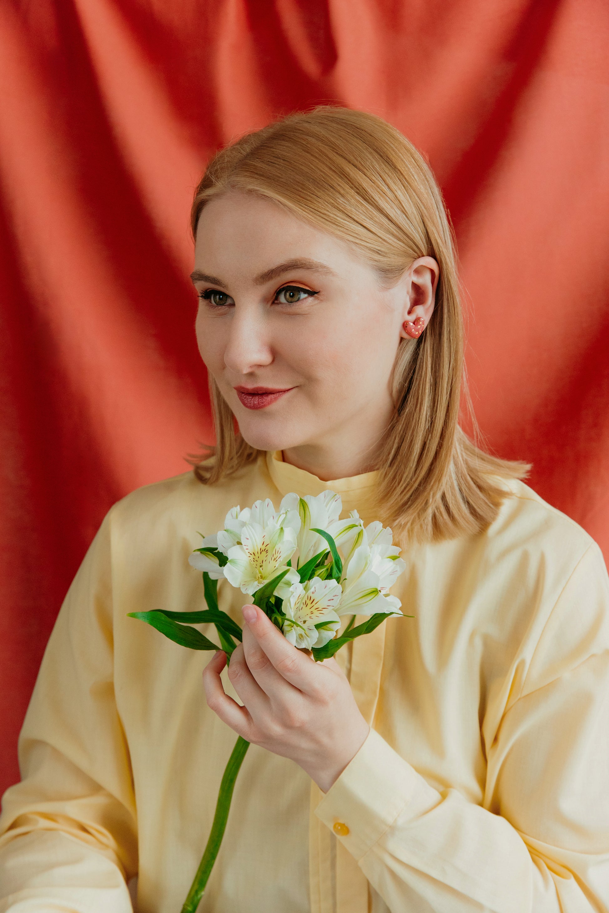 A woman with straight blonde hair, in a pale yellow shirt, holds white flowers and gently smiles before an orange-red backdrop, modeling Rozenthal Jewelry’s handmade Heart Studs porcelain earrings from Latvia.