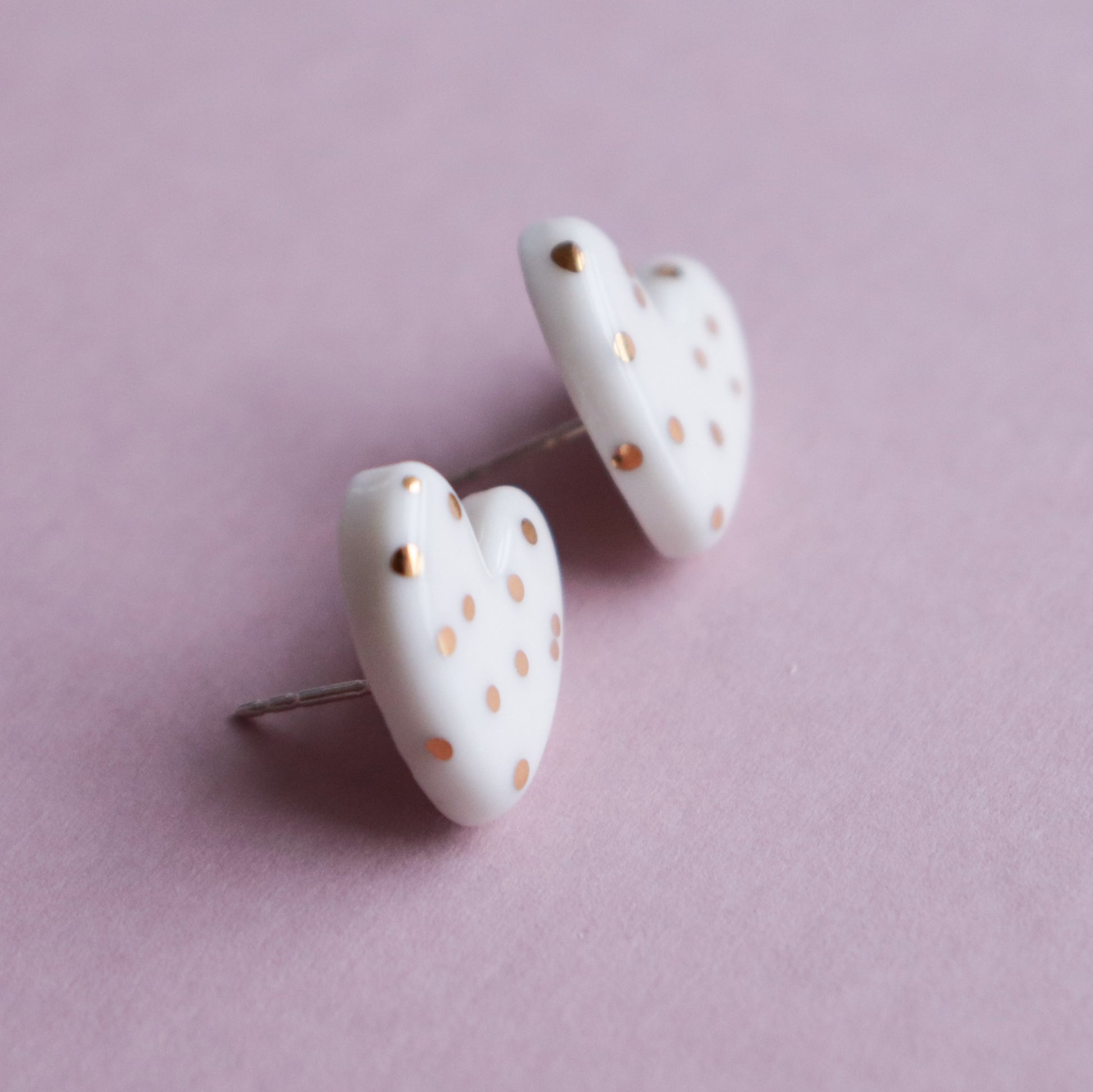 The Heart Studs by Rozenthal Jewelry are white, heart-shaped earrings adorned with small gold dots, handmade in Latvia and shown on a soft pink background.
