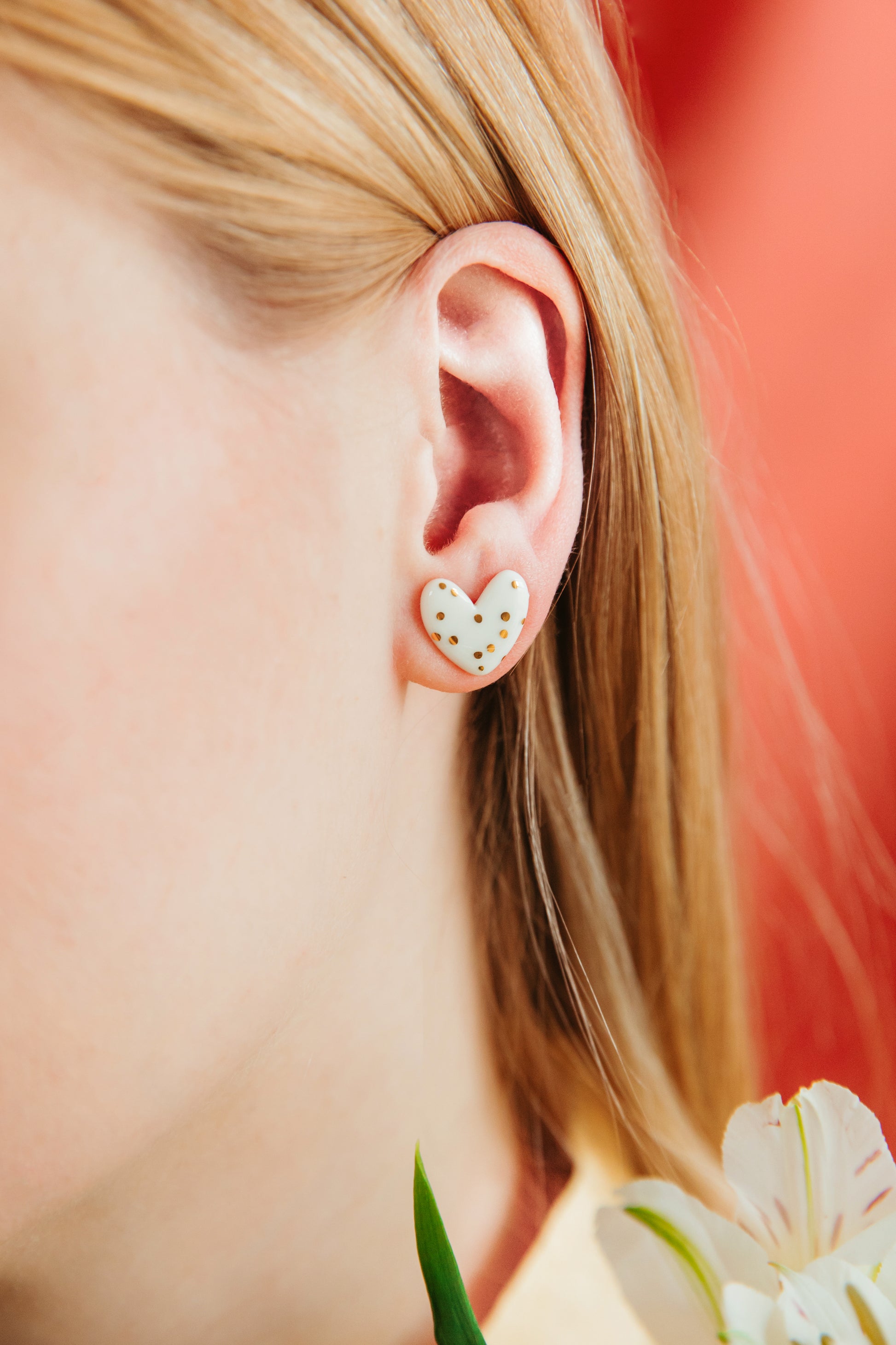 A close-up of a person with long blonde hair wearing Rozenthal Jewelry’s Heart Studs—white porcelain heart-shaped earrings adorned with gold dots. A white flower appears in the bottom right corner.