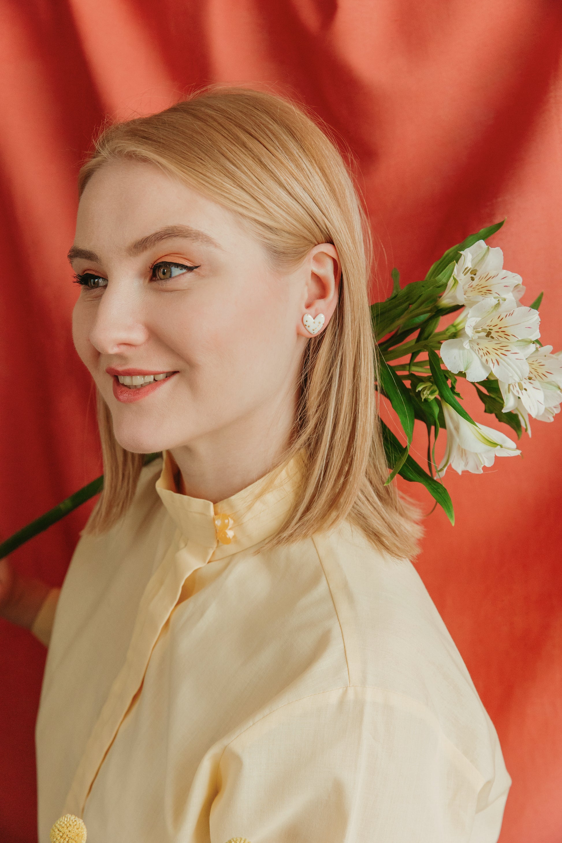 A smiling blonde woman in a pale yellow shirt wears Rozenthal Jewelrys Heart Studs earrings and holds white flowers over her shoulder, standing in front of a red fabric backdrop.