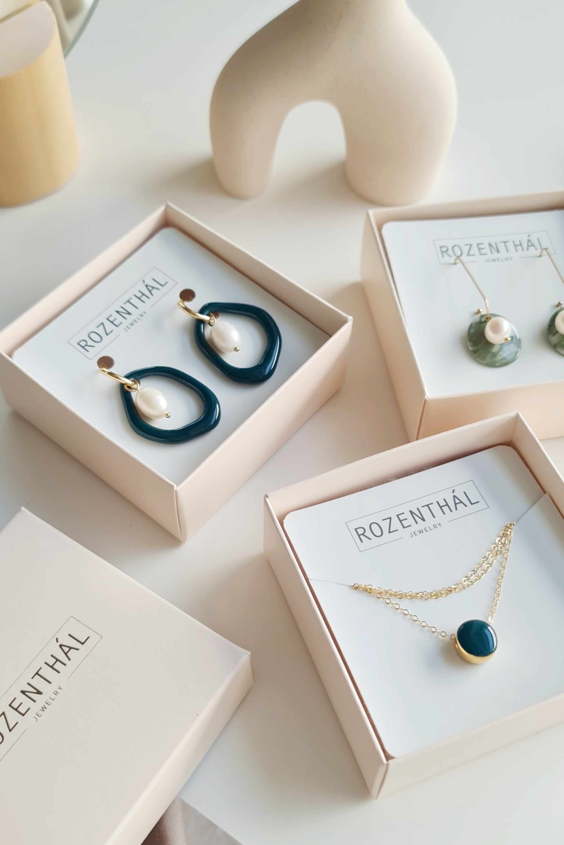 Three Rozenthal Jewelry gift boxes showcase elegant pieces: dark blue and pearl drop earrings, green and pearl drop earrings, and a gold necklace with a blue pendant—all stylishly presented in gift packaging on a light surface.