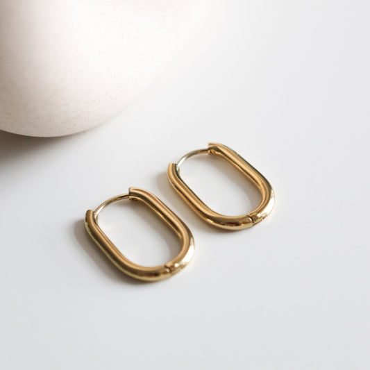 A pair of Rozenthal Jewelry Large Hoops in gold stainless steel are displayed on a white surface, with a smooth, cream-colored ceramic object partially visible in the background.