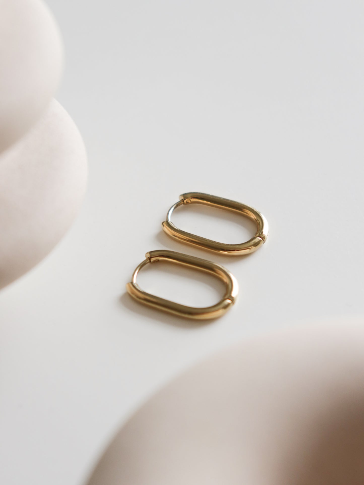 A pair of Large Hoops by Rozenthal Jewelry shown on a smooth white surface, with soft, out-of-focus beige shapes in the foreground and background.