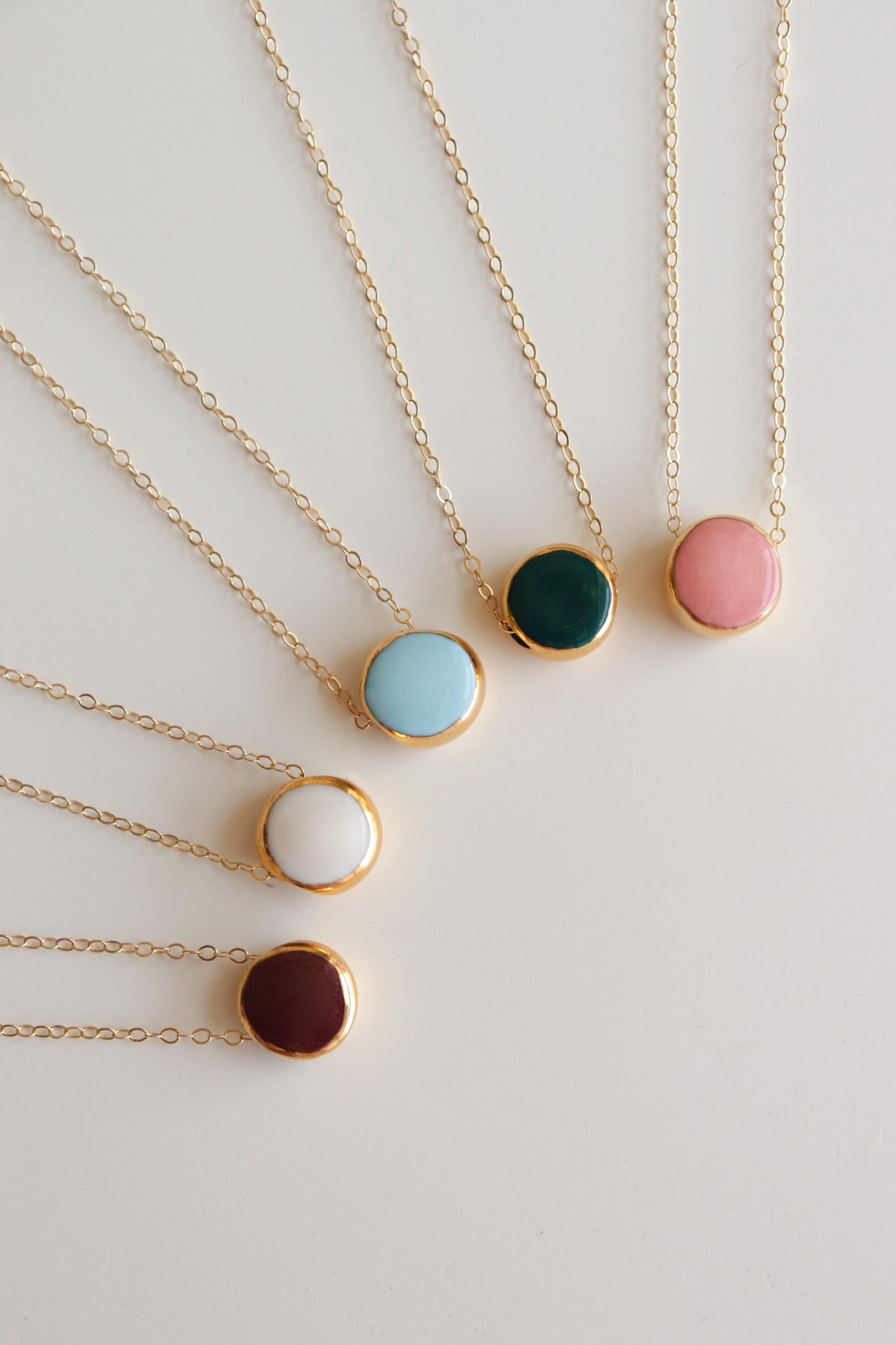 Six Rozenthal Jewelry Round Necklaces are arranged in a fan shape, each featuring a gold chain and a round porcelain pendant with centers in light blue, dark green, pink, white, brown, or turquoise.