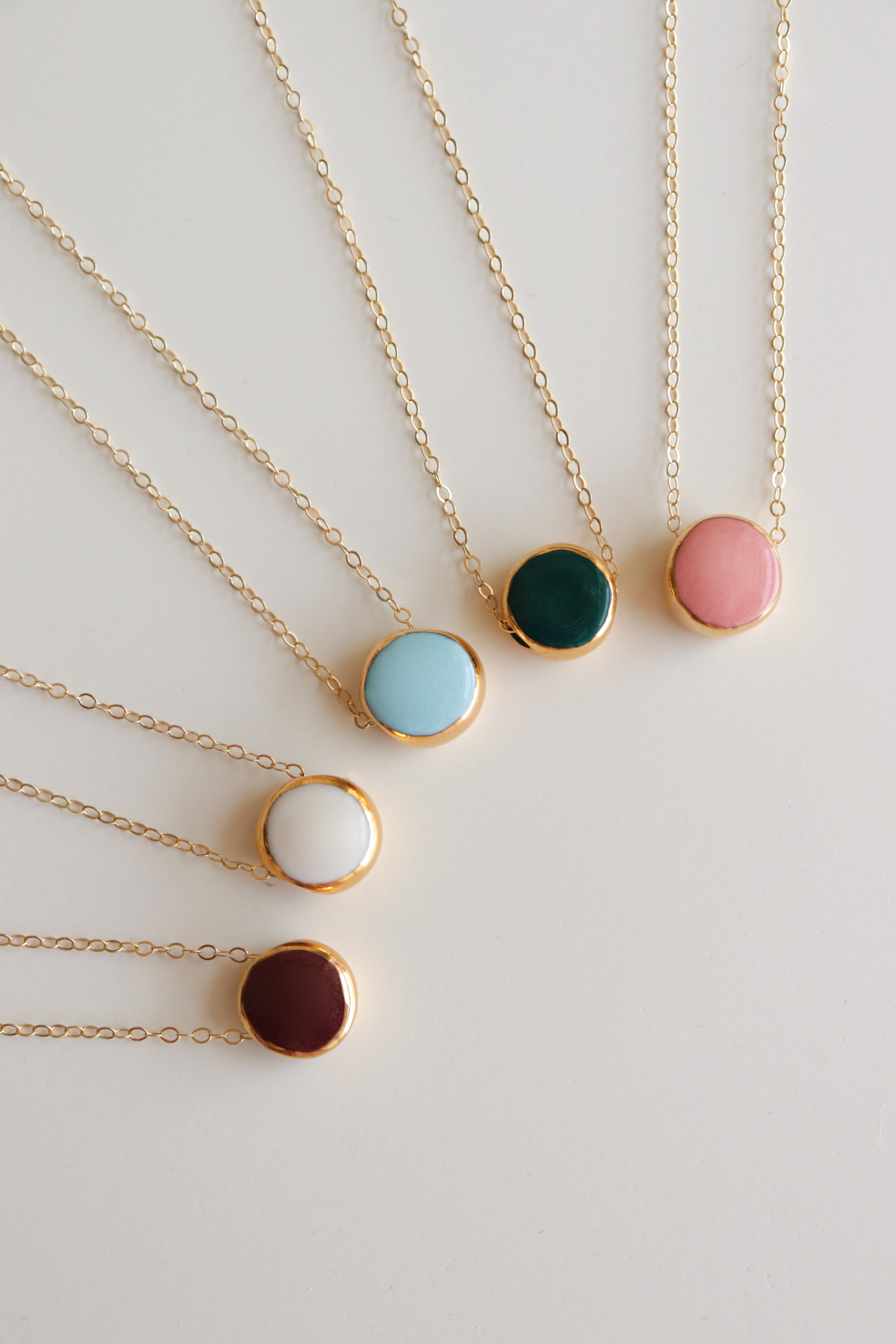 Six Rozenthal Jewelry Round Necklaces are arranged in a fan shape, each featuring a gold chain and a round porcelain pendant with centers in light blue, dark green, pink, white, brown, or turquoise.