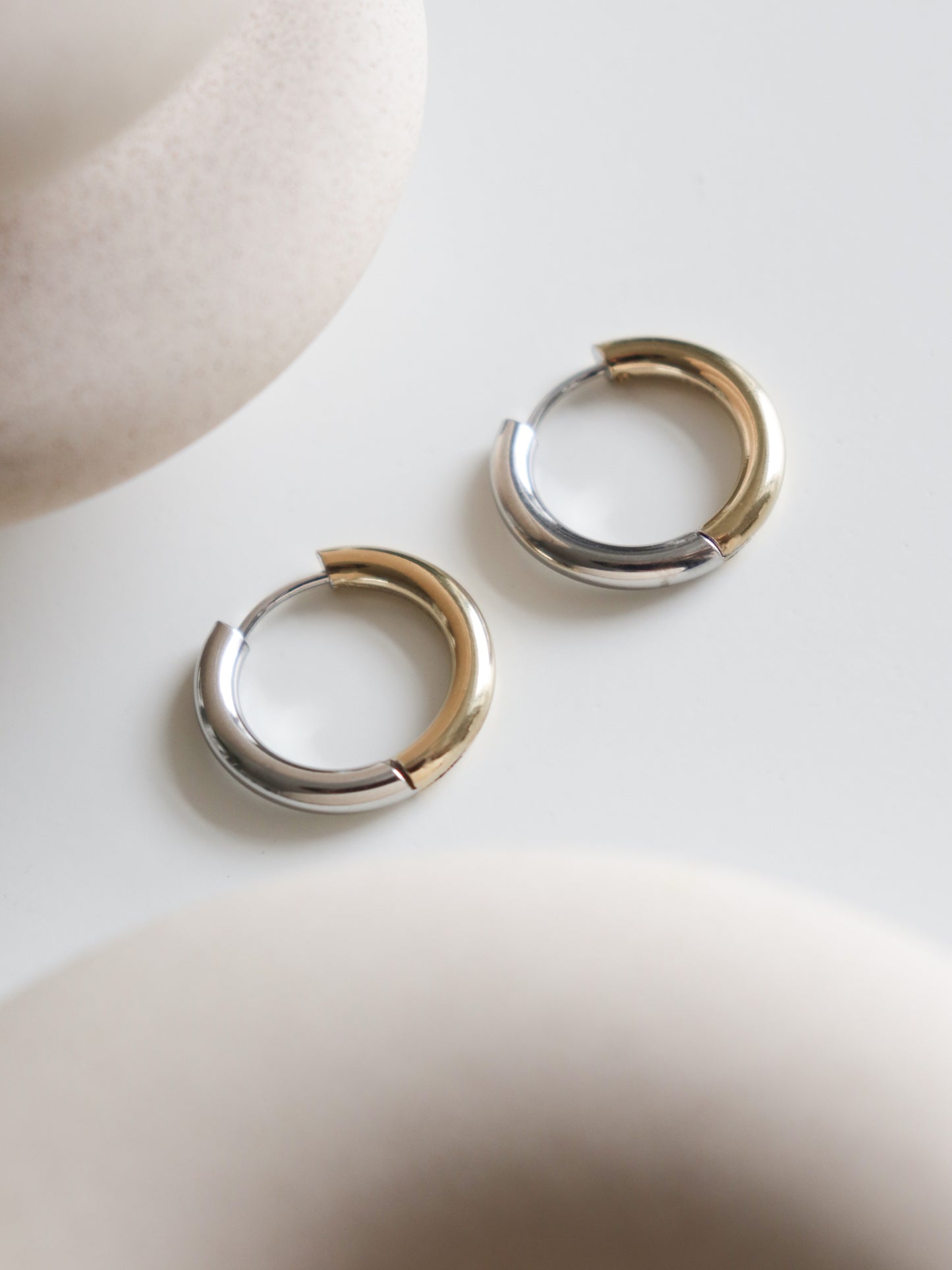 Two Gold and Silver Hoops by Rozenthal Jewelry rest on a smooth white surface, with blurred round beige objects in the foreground and background.
