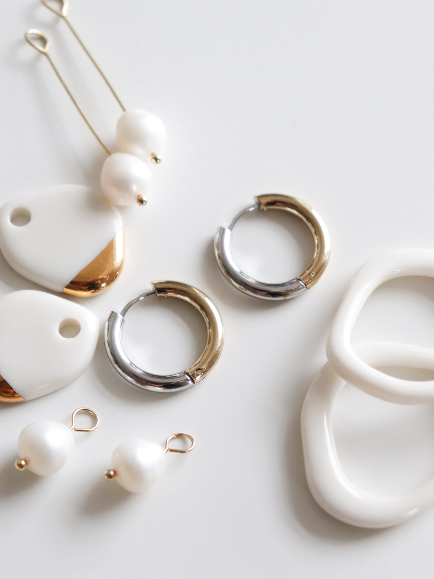 A flat lay of Rozenthal Jewelry’s Gold and Silver Hoops is displayed on a white surface, alongside enamel pendants, pearl charms, and white irregular-shaped rings.