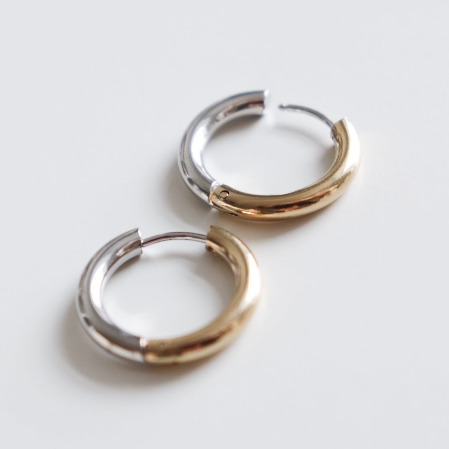 Two Gold and Silver Hoops by Rozenthal Jewelry rest side by side on a white surface; each earring features a smooth, shiny finish with half silver and half gold tones.