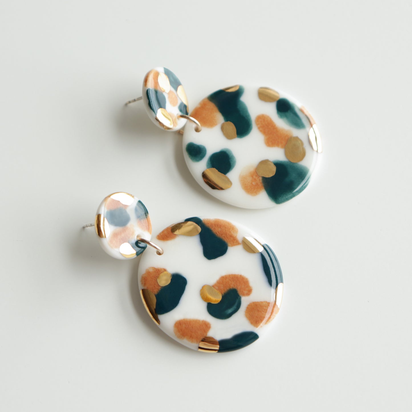 The Tache Earrings by Rozenthal Jewelry are round, dangling porcelain earrings with a white base and abstract teal, mustard yellow, and rust orange spots. These glossy, hypoallergenic pieces are shown on a plain white background.