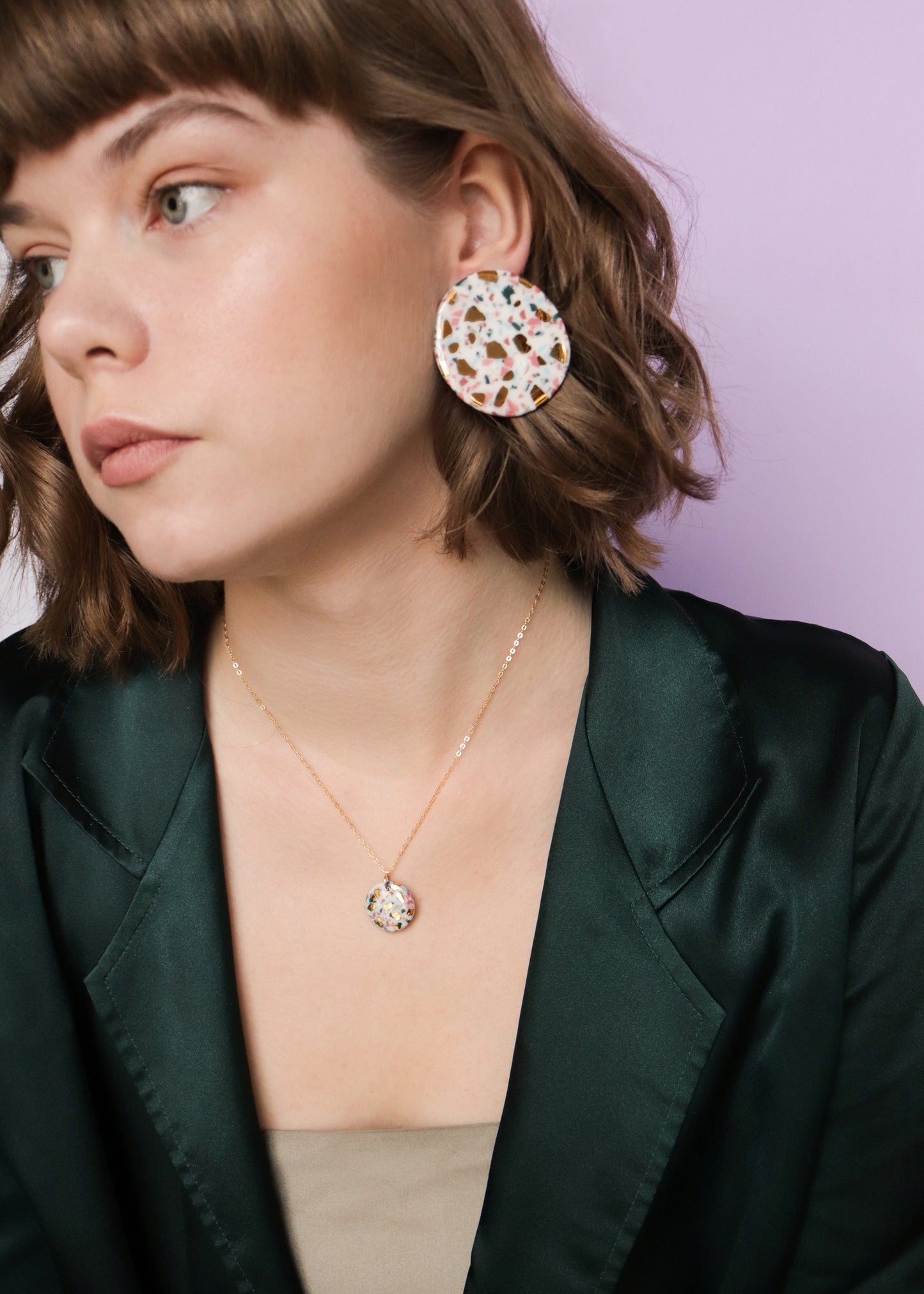 A woman with short brown hair wears a dark green blazer, beige top, the Astilla Recycled Necklace by Rozenthal Jewelry, and large terrazzo-style earrings. Against a lavender background, she looks to the side, showcasing her love for handmade jewelry.