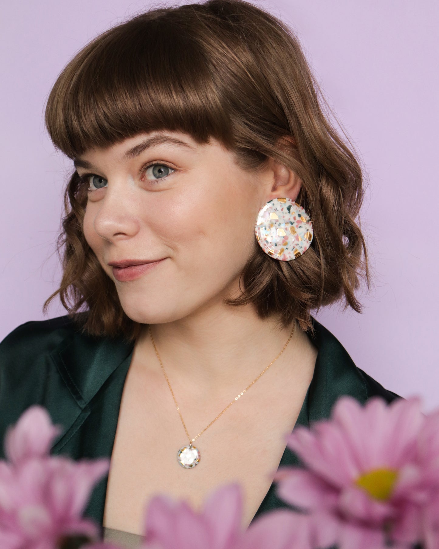 A young woman with short brown hair and bangs smiles gently, wearing a dark green jacket, a colorful round earring, and the Astilla Recycled Necklace by Rozenthal Jewelry. Pink flowers are in front of her against a light purple background.