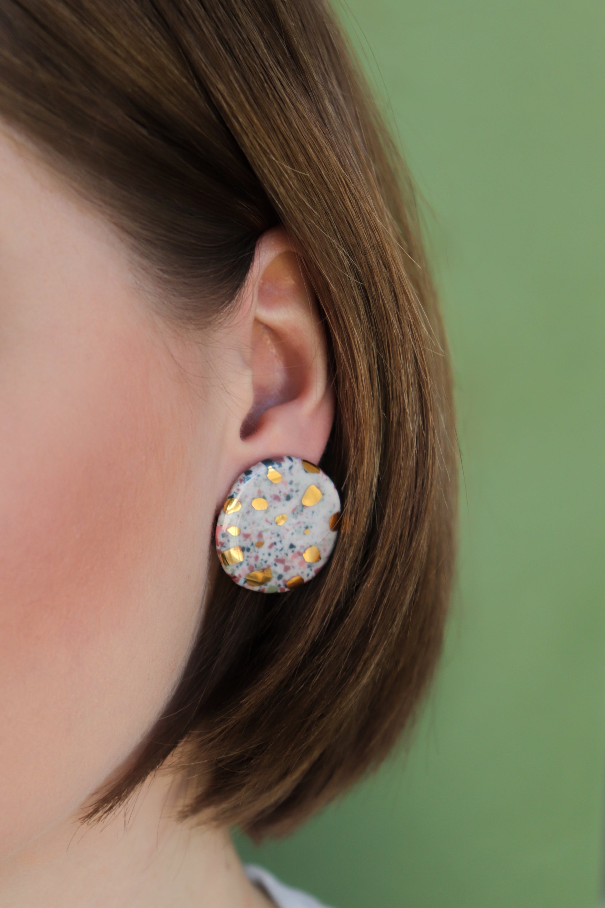 Close-up of a person with short brown hair wearing Astilla Recycled Studs / M by Rozenthal Jewelry—large round porcelain earrings with a terrazzo-style speckled design, set against a solid light green background.