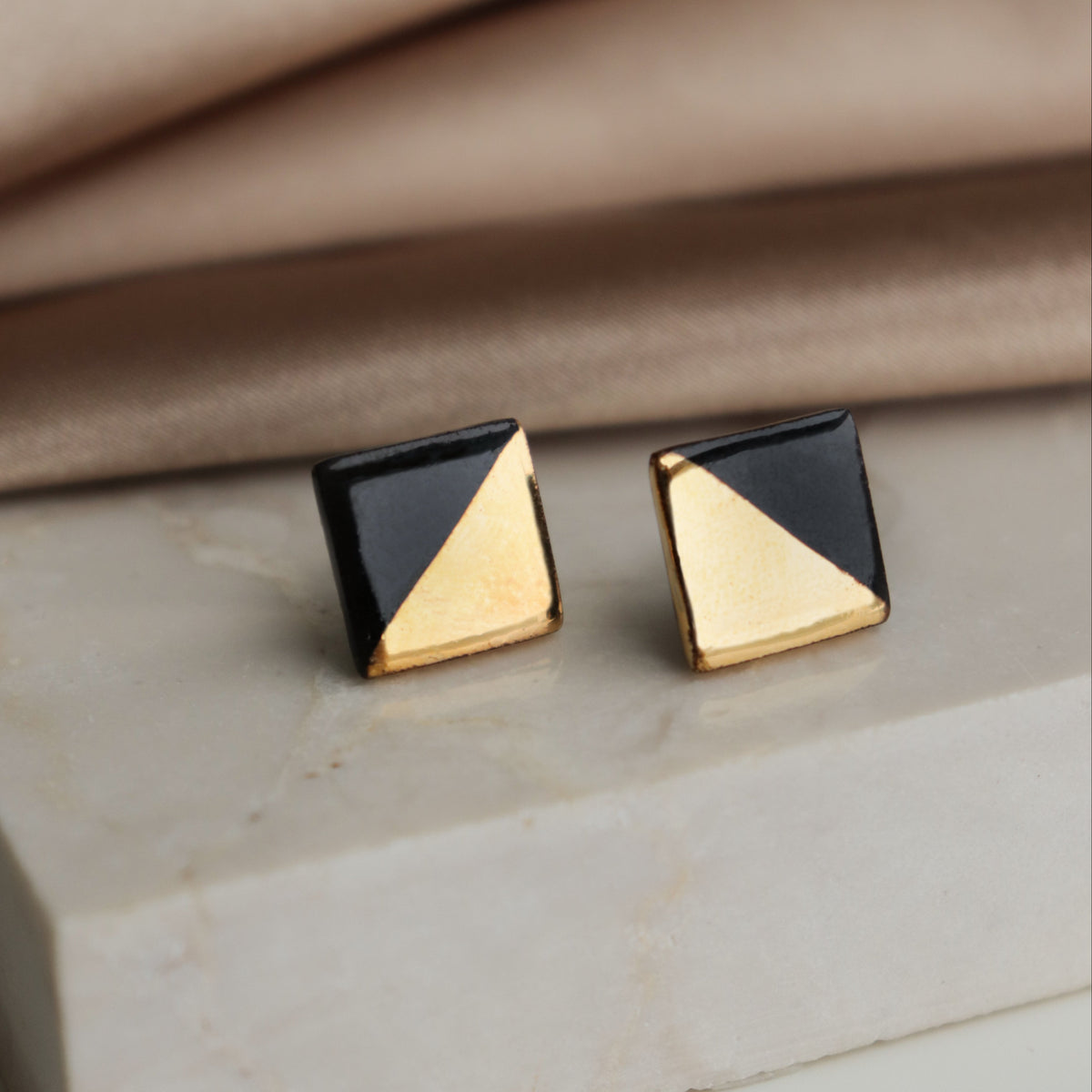 A pair of geometric square stud earrings with a split black and gold triangular design, displayed on a light marble block with a beige fabric background.