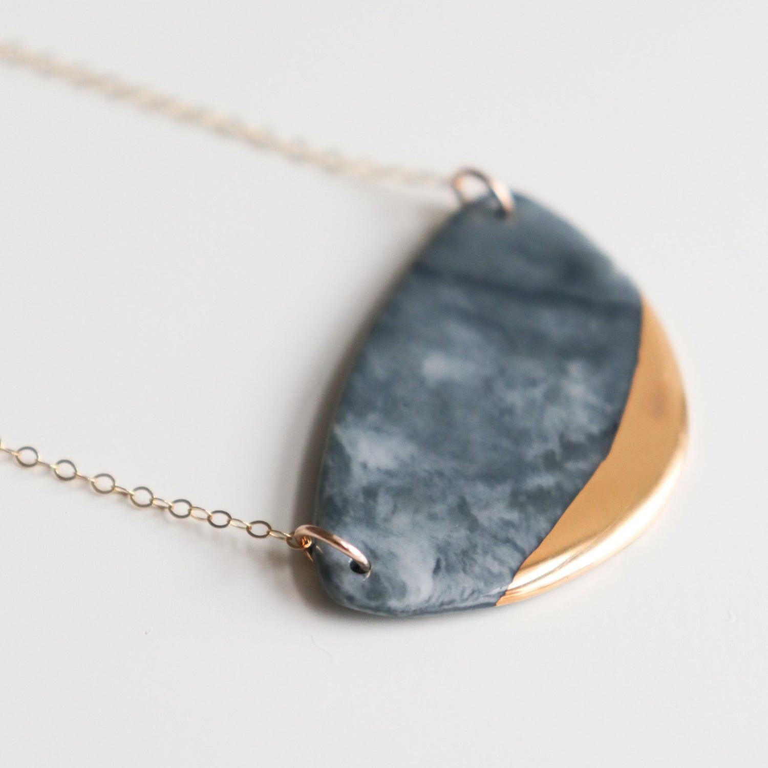 The Leaf Necklace by Rozenthal Jewelry features a semi-circular porcelain pendant with a marbled gray and gold design on a delicate gold chain—an elegant, handmade piece for jewelry lovers.