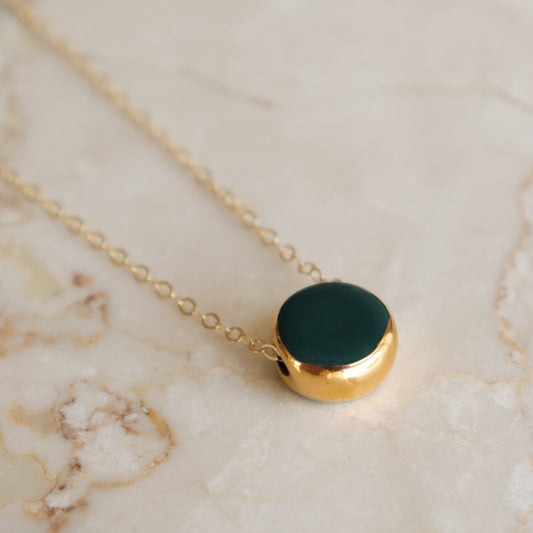 A gold chain necklace with a round gold pendant featuring a dark green inlay, displayed on a light beige marble surface.