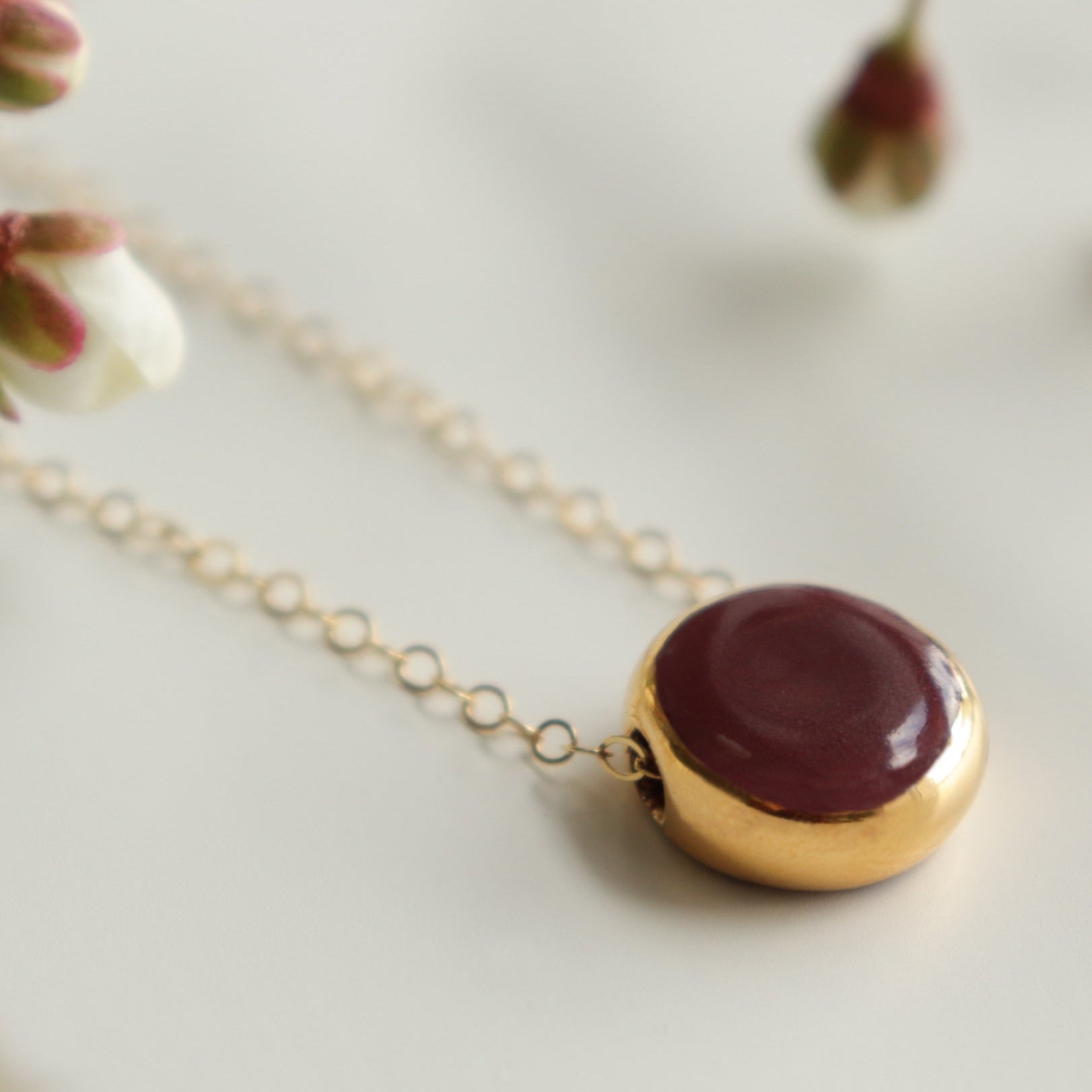 A gold chain necklace with a round pendant featuring a deep burgundy red center and a gold rim, styled on a light surface with soft-focus white blossoms.