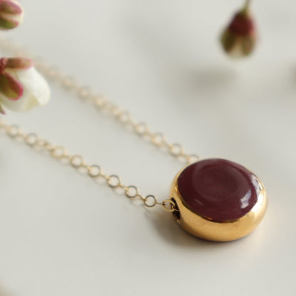 A gold chain necklace with a round pendant featuring a deep burgundy red center and a gold rim, styled on a light surface with soft-focus white blossoms.