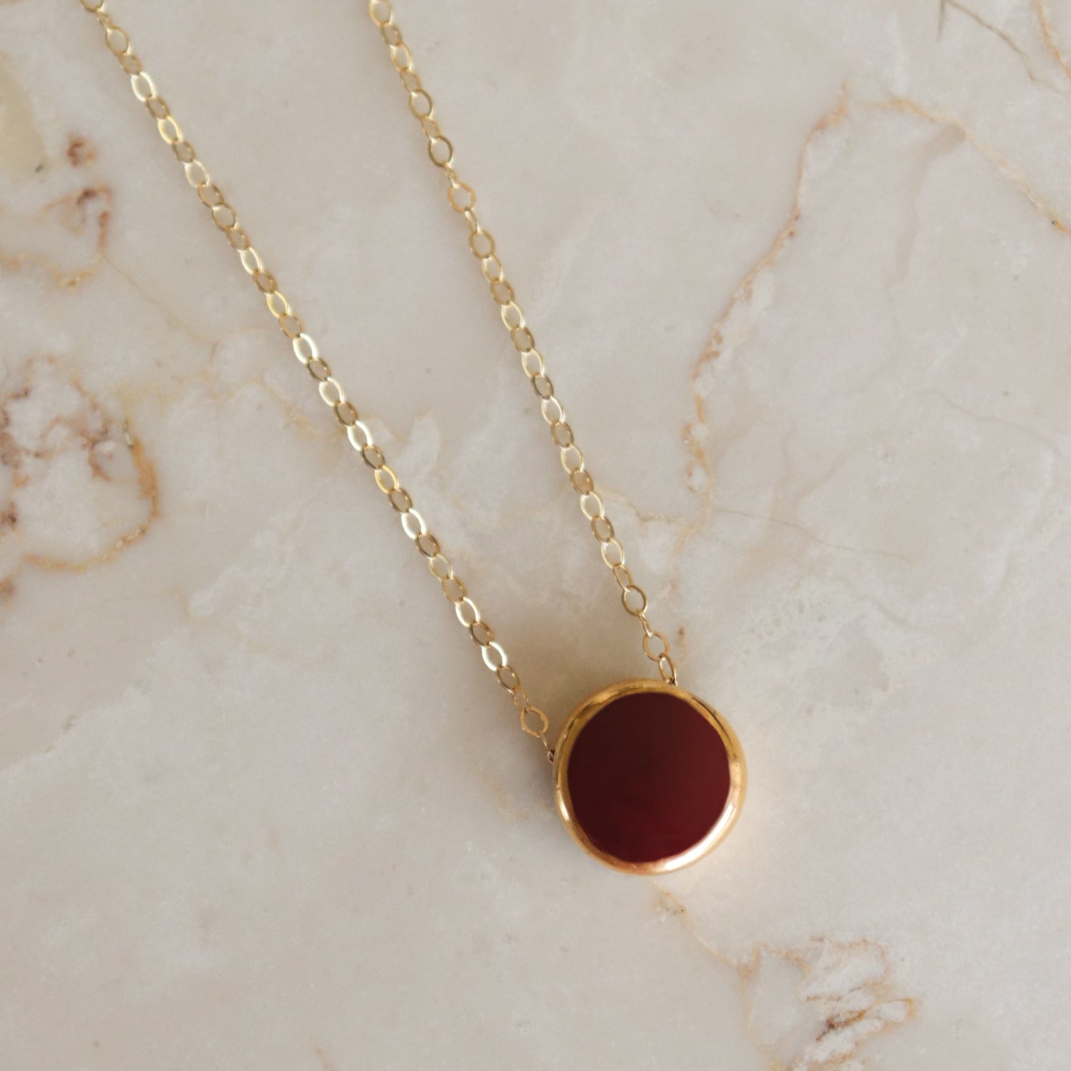 The Rozenthal Jewelry Round Necklace features a gold chain with a deep red centered round pendant, showcased on light marble. Ideal for those who love minimalist jewelry with timeless style.