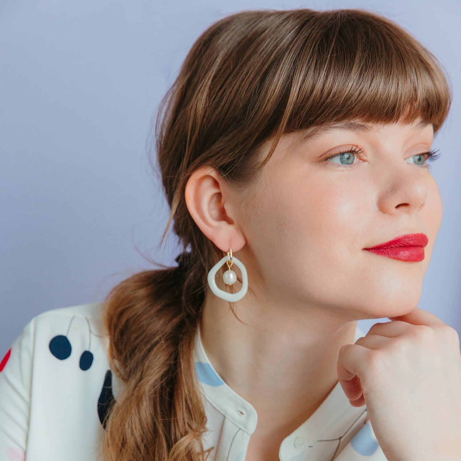 A woman with long brown hair in a ponytail, wearing a white blouse with colorful spots and Rozenthal Jewelrys Arp Pearl Hoops, smiles gently and rests her chin on her hand against a light purple background.