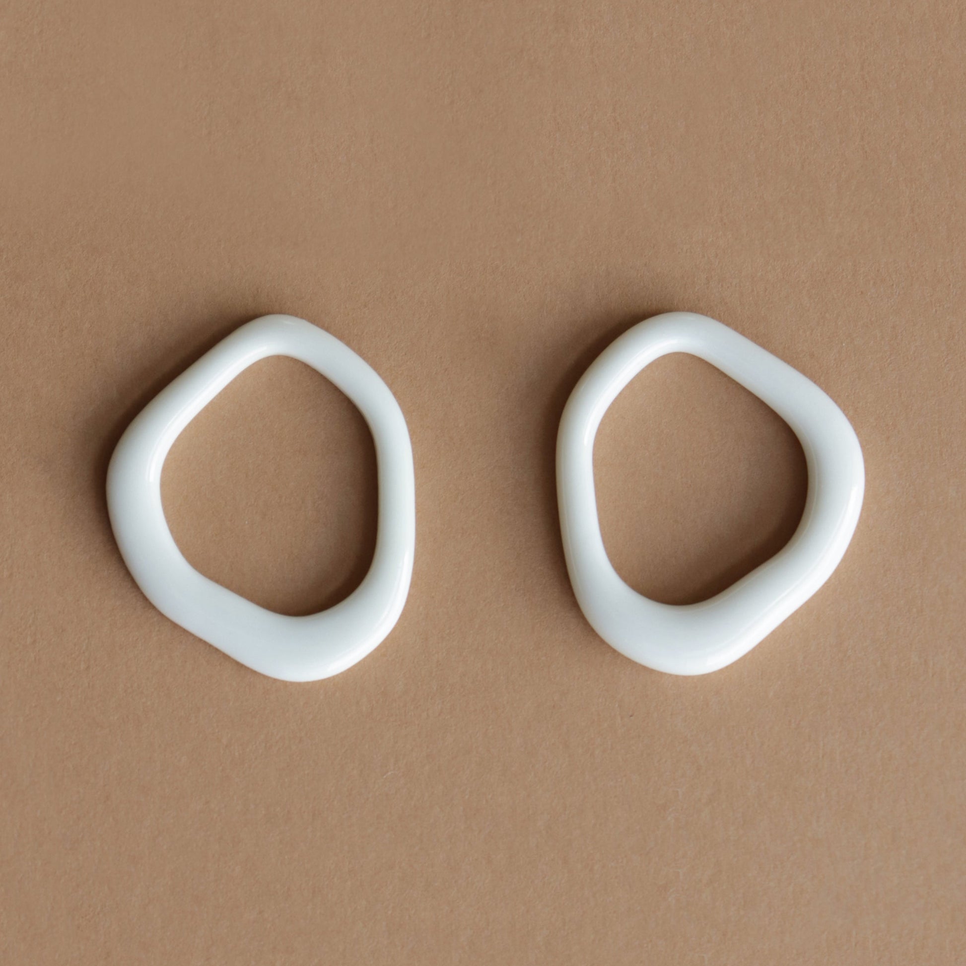 The Arp Pearl Hoops by Rozenthal Jewelry feature two irregular, white, oval ceramic rings with smooth edges and organic, asymmetrical forms, highlighting the unique charm of handmade jewelry on a light brown surface.