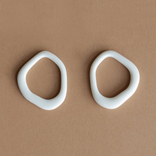 The Arp Pearl Hoops by Rozenthal Jewelry feature two irregular, white, oval ceramic rings with smooth edges and organic, asymmetrical forms, highlighting the unique charm of handmade jewelry on a light brown surface.