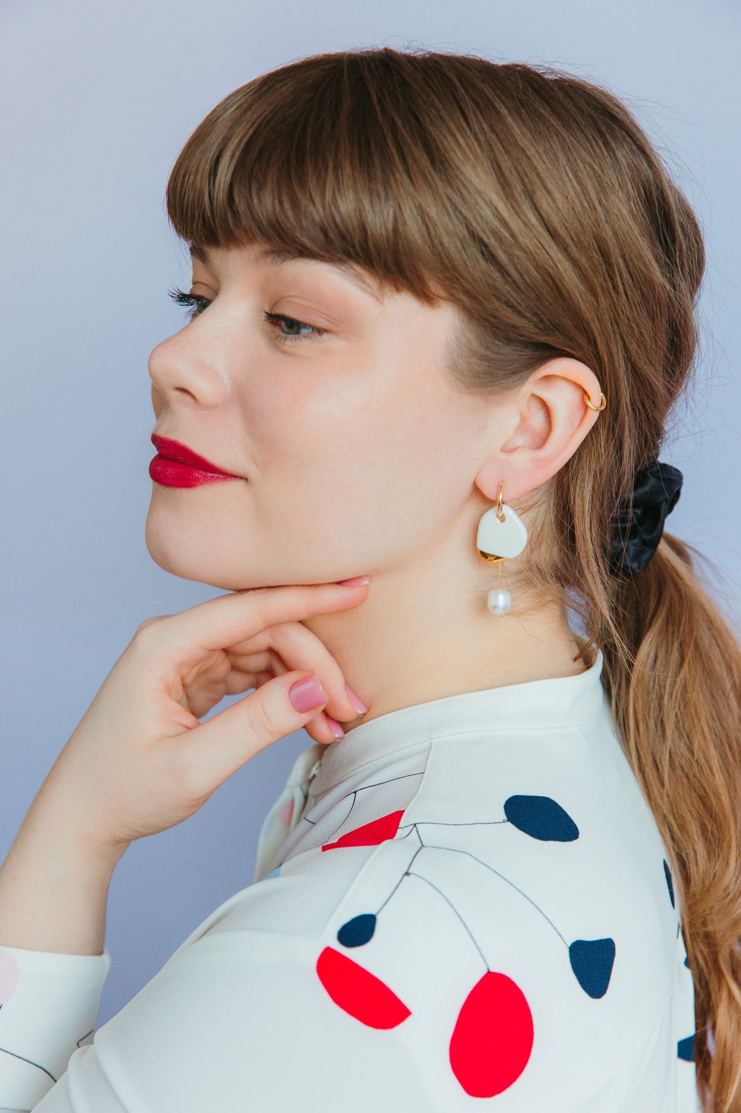 A woman with light brown hair in a ponytail wears red lipstick and Rozenthal Jewelrys Arp Pearl Hoops. She poses in a white top with red and navy patterns, hand under her chin, against a light purple background.