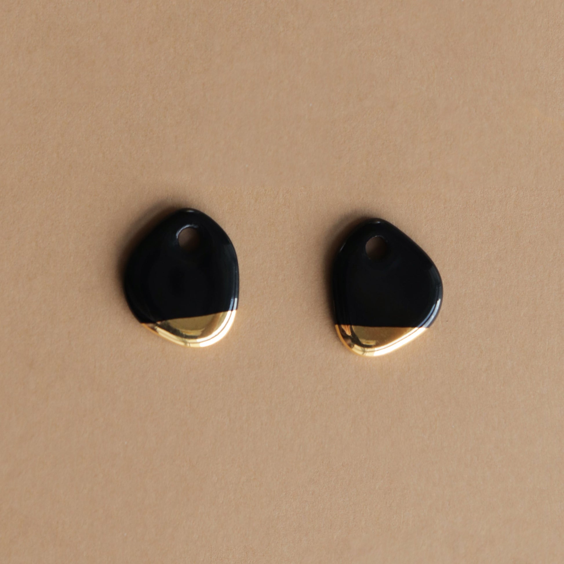 The Arp Pearl Hoops by Rozenthal Jewelry feature two irregularly shaped black and gold charms with a small top hole, ideal for handmade jewelry, shown on a light brown surface.