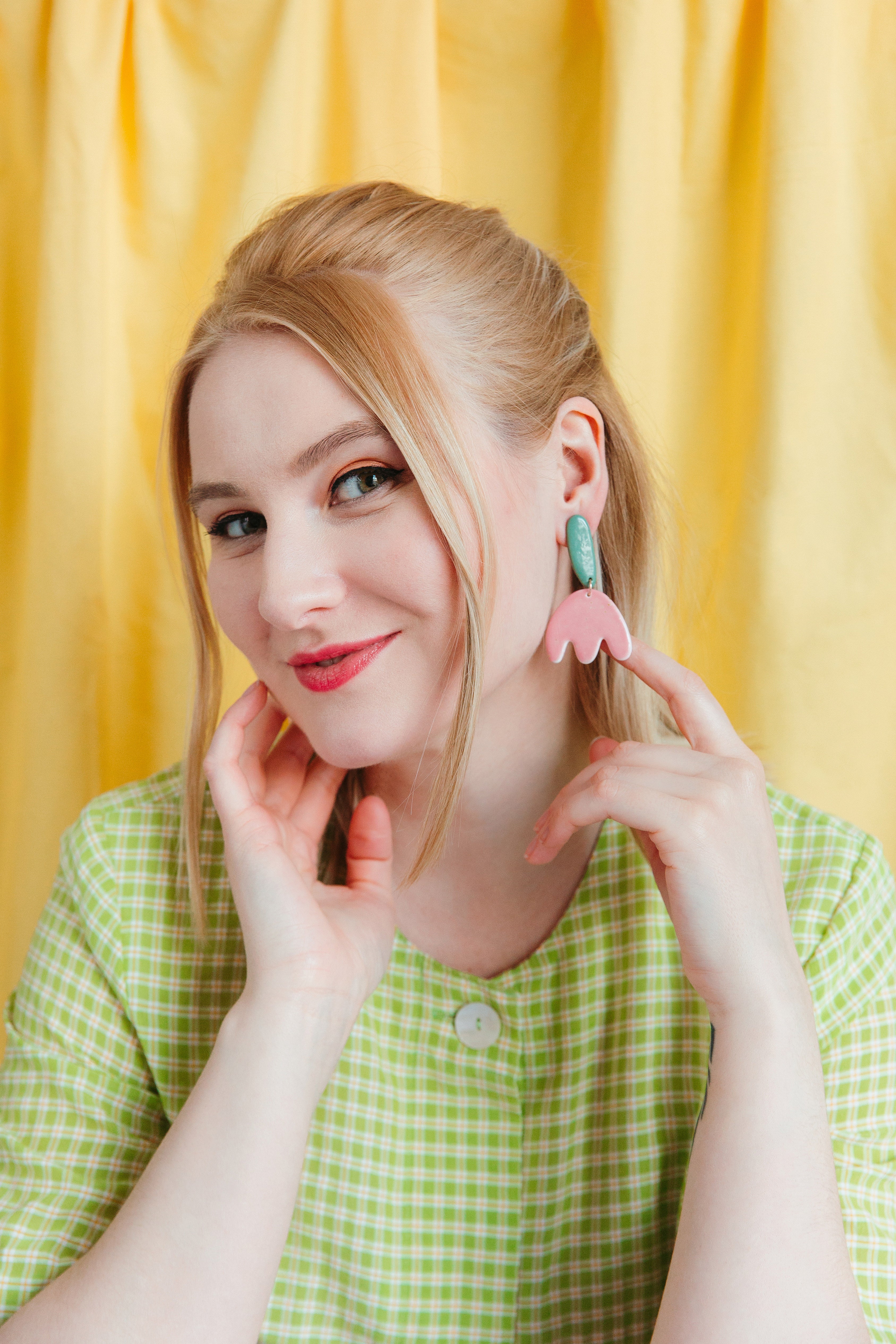 A smiling blonde woman in a green checkered dress points to her handmade Tulip Earrings by Rozenthal Jewelry, featuring pink and green flowers, while standing in front of a yellow curtain.