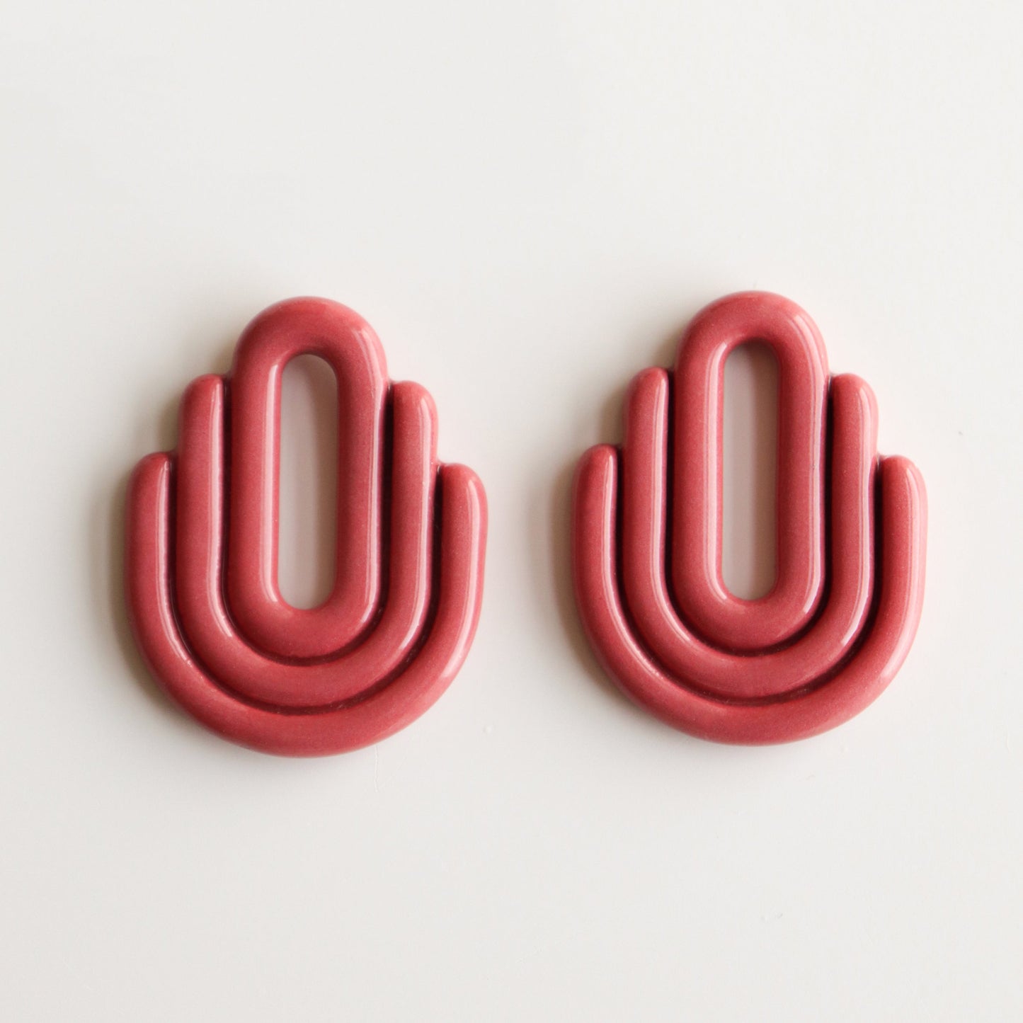 Two pink, geometric Art Deco Earrings from Rozenthal Jewelry, each with three rounded tiers and an open oval center, sit side by side on a white background; their arch-shaped design resembles a stylized hand or rainbow.