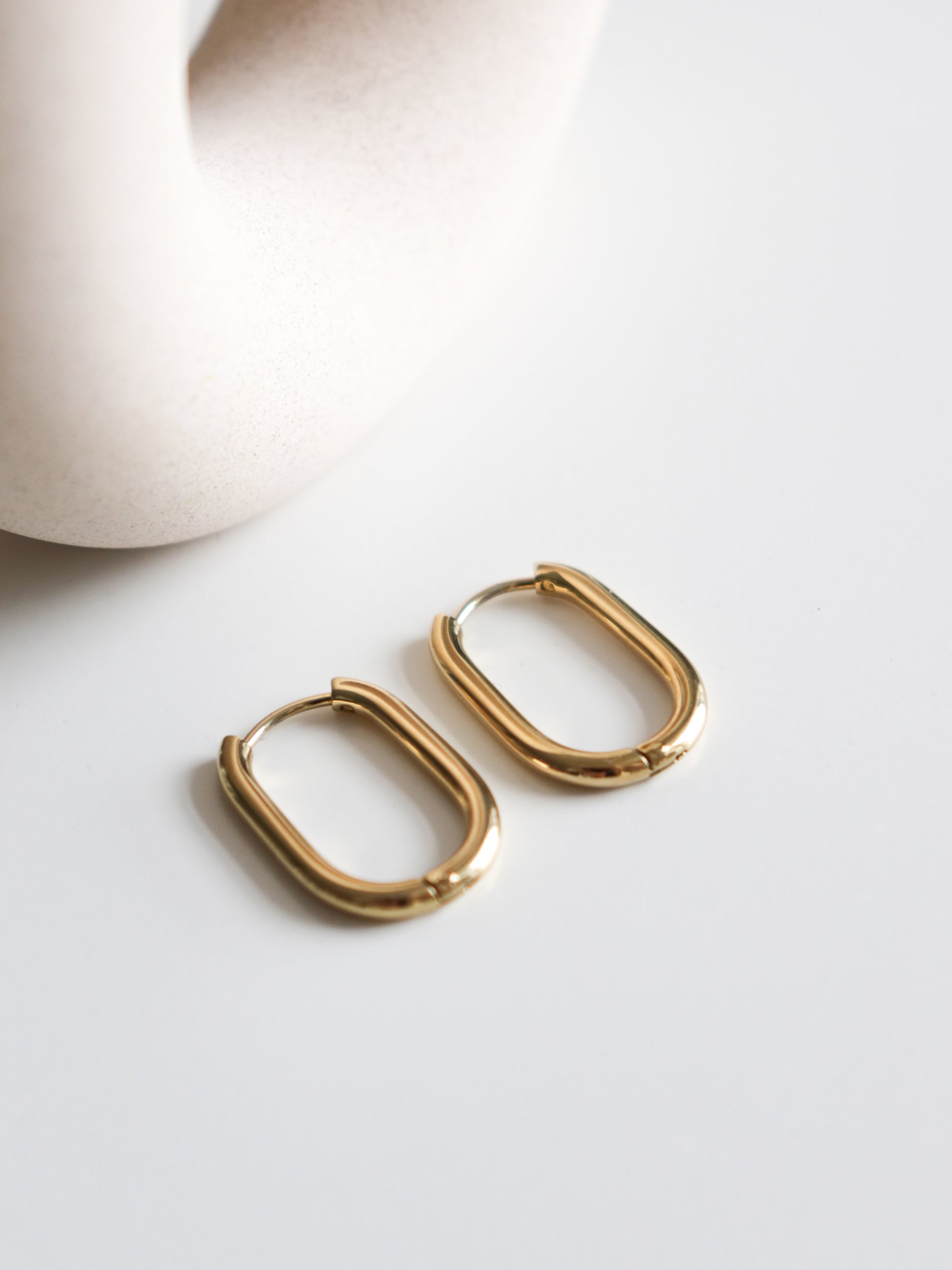 A pair of Rozenthal Jewelry Large Hoops in gold stainless steel are displayed on a white surface, with a smooth, cream-colored ceramic object partially visible in the background.