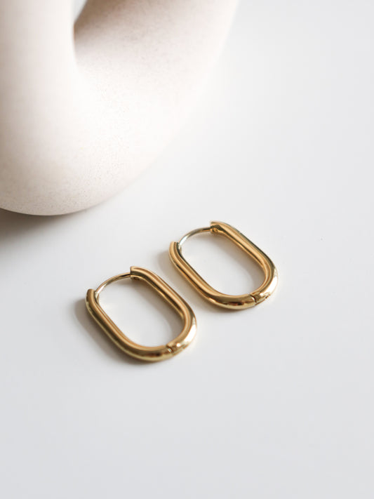 A pair of Rozenthal Jewelry Large Hoops in gold stainless steel are displayed on a white surface, with a smooth, cream-colored ceramic object partially visible in the background.